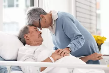 Eine Frau drückt ihren Kopf einfühlsam an den Kopf eines älteren Mannes, der in einem Krankenhausbett einer Palliativstation liegt.