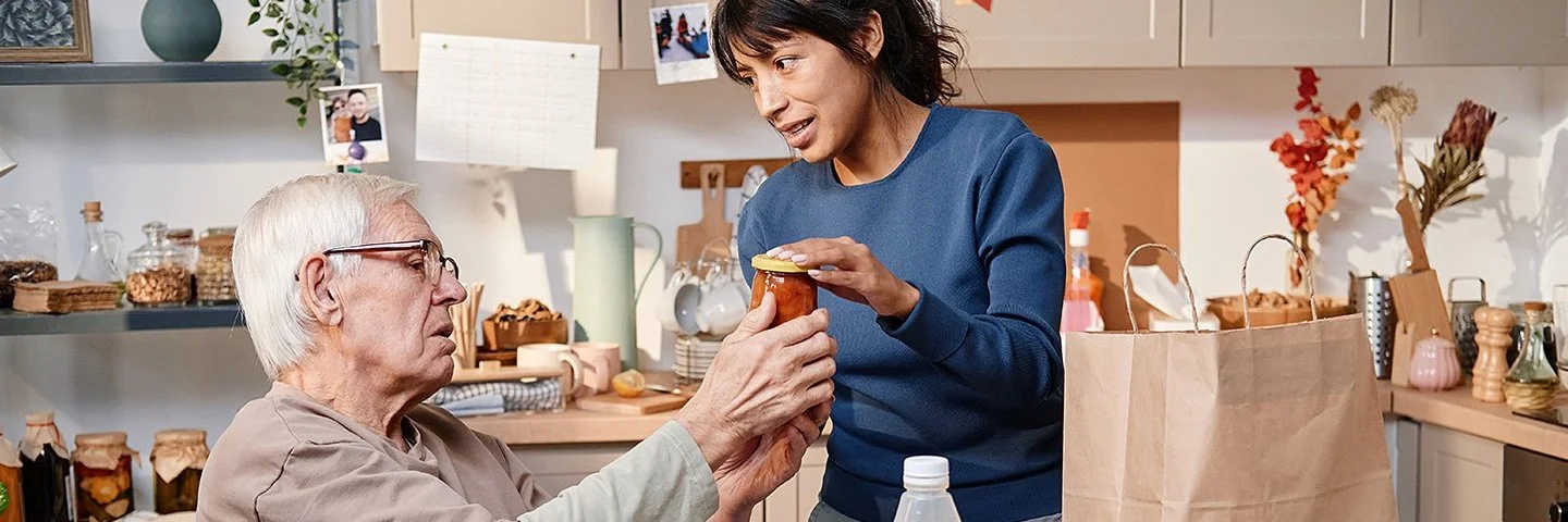 Eine Frau hat Lebensmittel für einen älteren Mann im Rollstuhl gekauft. Beide befinden sich in einer Küche. Die Frau zeigt dem Mann die eingekauften Lebensmittel. Auf dem Esstisch stehen eine Einkaufstüte und verschiedene Lebensmittel wie Eier, Saft und Obst. Im Hintergrund sind auf der Küchenanrichte und in Regalen verschiedene Küchenutensilien zu sehen.