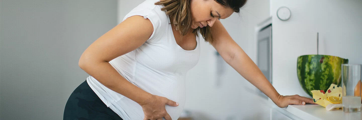 Eine Frau mit Wehen stützt sich auf der Küchenzeile auf und hält sich den Babybauch.