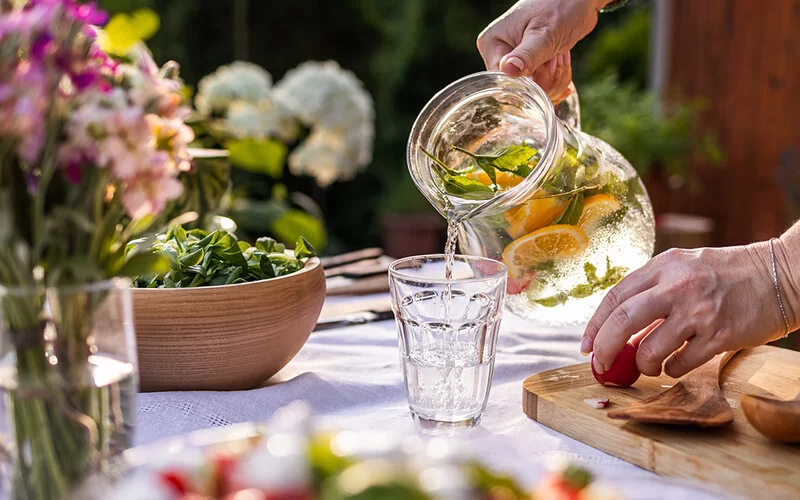 Eine ältere Frau gießt aus einer Karaffe mit Zitrone und Minze aromatisiertes Wasser in ein Glas, das auf einem gedeckten Tisch steht.