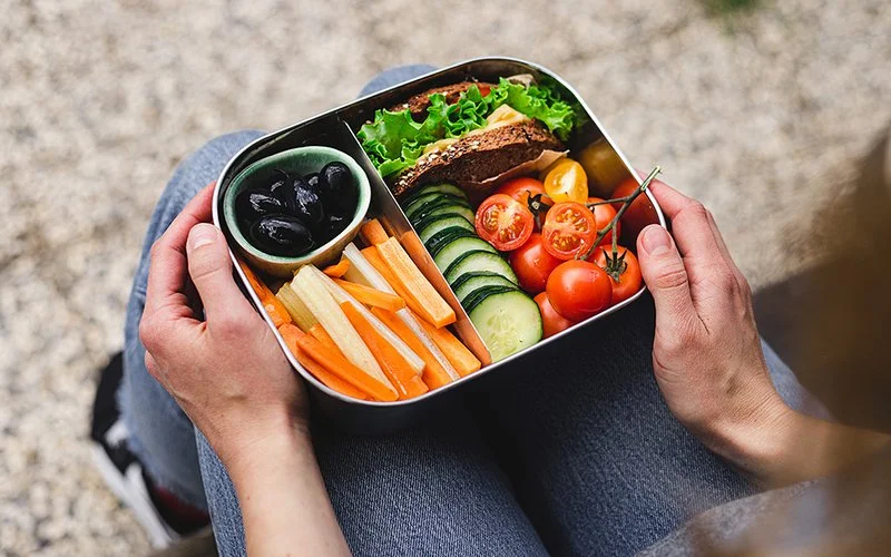 Eine Lunchbox auf den Knien einer Frau. In der Box befinden sich Karotten- und Sellerieschnitze, Oliven, Gurkenscheiben, Cherrytomaten und ein mit Salat und Käse belegtes Vollkornbrot.