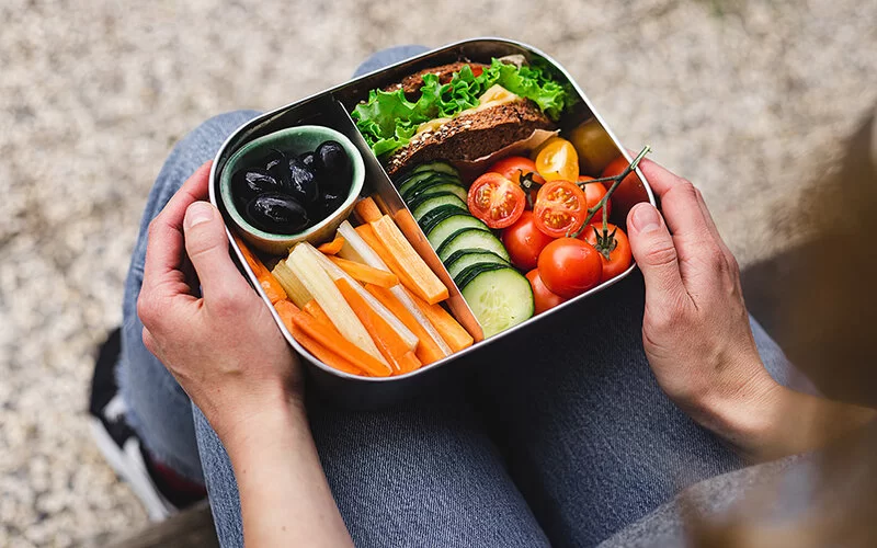 Eine Lunchbox auf den Knien einer Frau. In der Box befinden sich Karotten- und Sellerieschnitze, Oliven, Gurkenscheiben, Cherrytomaten und ein mit Salat und Käse belegtes Vollkornbrot.