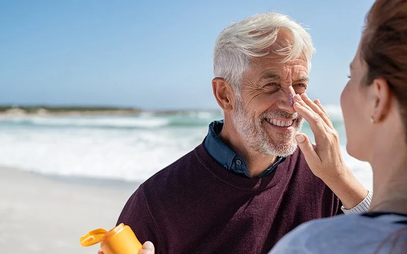 Ein älterer Herr und eine Frau sind am Strand. Sie cremt ihm die Nase mit Sonnenschutz ein.