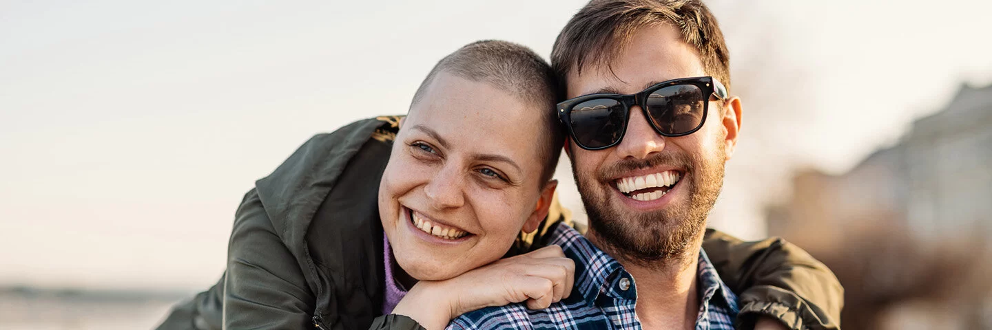 Eine junge Frau mit kurzgeschorenen Haaren und ein junger Mann mit Sonnenbrille stehen am Strand und lächeln in die Kamera.