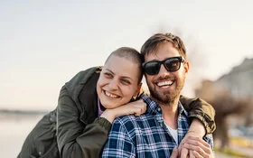 Eine junge Frau mit kurzgeschorenen Haaren und ein junger Mann mit Sonnenbrille stehen am Strand und lächeln in die Kamera.
