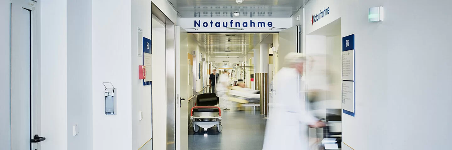 Ein langer, heller Krankenhausflur mit einem Schild auf dem „Notaufnahme" steht. Eine Person in medizinischer Kleidung eilt nach rechts. Im Hintergrund sind weitere Personen und ein Krankenbett zu erkennen, der gesamte Bereich wirkt geschäftig.