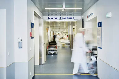 Ein langer, heller Krankenhausflur mit einem Schild auf dem „Notaufnahme" steht. Eine Person in medizinischer Kleidung eilt nach rechts. Im Hintergrund sind weitere Personen und ein Krankenbett zu erkennen, der gesamte Bereich wirkt geschäftig.
