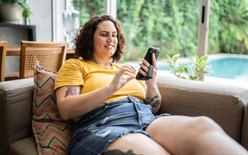 Eine junge mehrgewichtige Frau liegt bequem auf der Couch und surft mit ihrem Smartphone im Internet.