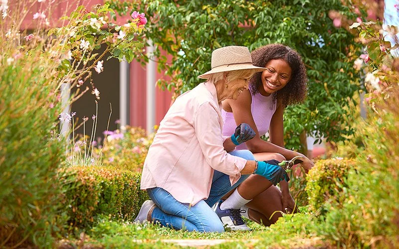 Zwei lächelnde Frauen knien draußen und machen Gartenarbeit.