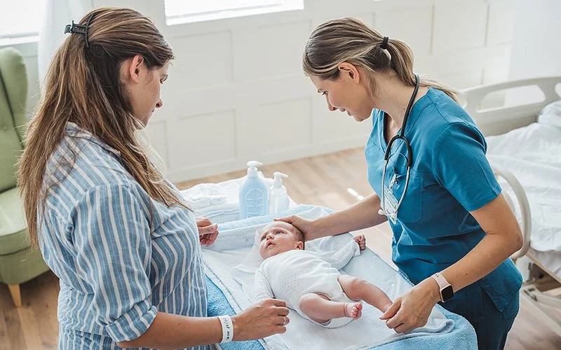 Ein Baby liegt auf dem Bett und schaut zur Mutter. Eine Pflegefachperson zeigt der Mutter, wie sie sich um das Baby kümmern kann.