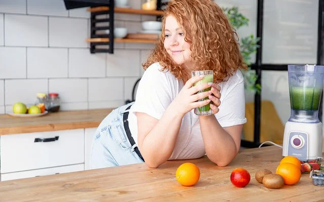 Eine junge Frau lehnt in einer Küche an der Arbeitsfläche und hält ein Glas grünen Smoothie in den Händen.