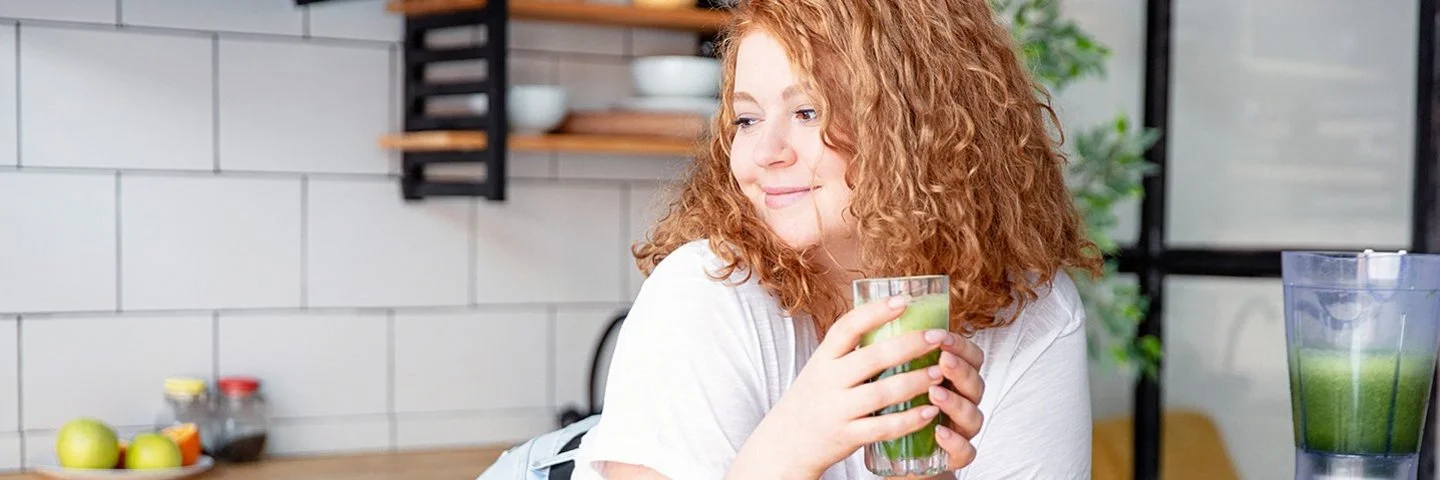 Eine junge Frau lehnt in einer Küche an der Arbeitsfläche und hält ein Glas grünen Smoothie in den Händen.