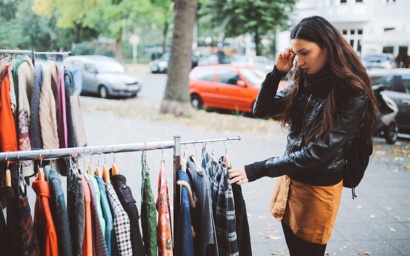 Eine junge Frau schaut sich Kleidung auf einem Ständer vor einem Secondhand-Shop an.