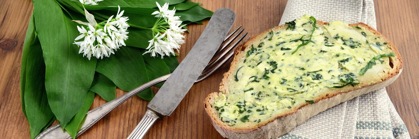 Eine mit Bärlauchbutter bestrichene Scheibe Brot liegt neben Bärlauchblättern und Silberbesteck auf einem Tisch.