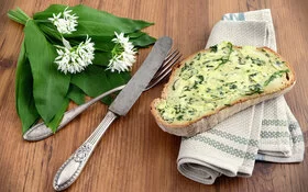 Eine mit Bärlauchbutter bestrichene Scheibe Brot liegt neben Bärlauchblättern und Silberbesteck auf einem Tisch.