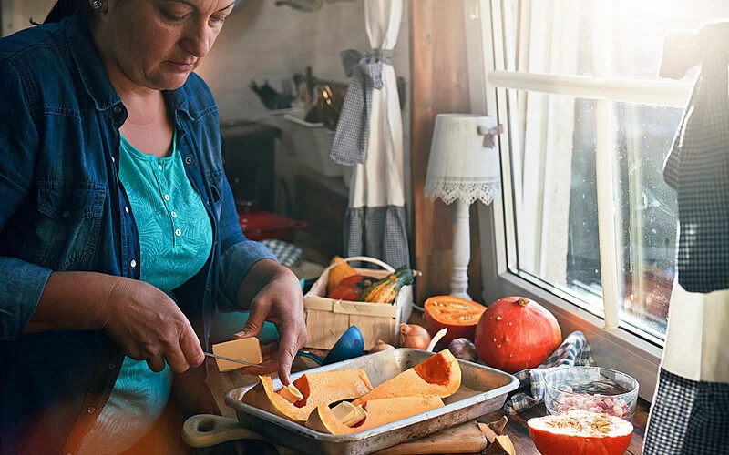 Eine Frau steht am Küchenfenster und befüllt eine Auflaufform mit Kürbisstücken.