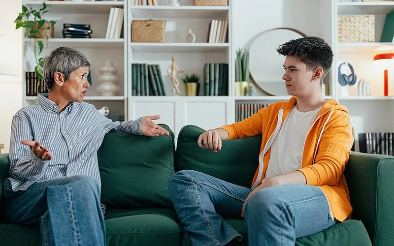 Auf einem Sofa in einem Wohnzimmer sitzen eine Frau und ein männlicher Teenager. Sie schauen sich in die Augen. Die Frau gestikuliert mit den Armen und spricht zu dem Jungen.