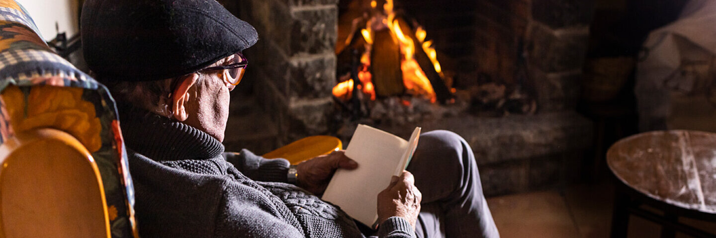 Ein älterer Mann sitzt lesend vor dem brennenden Kamin.