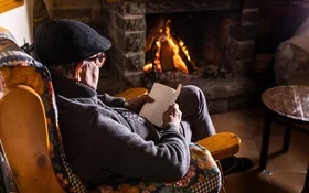 Ein älterer Mann sitzt lesend vor dem brennenden Kamin.