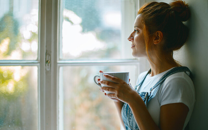 Eine junge Frau schaut lächelnd mit einer Tasse in der Hand aus dem Fenster.
