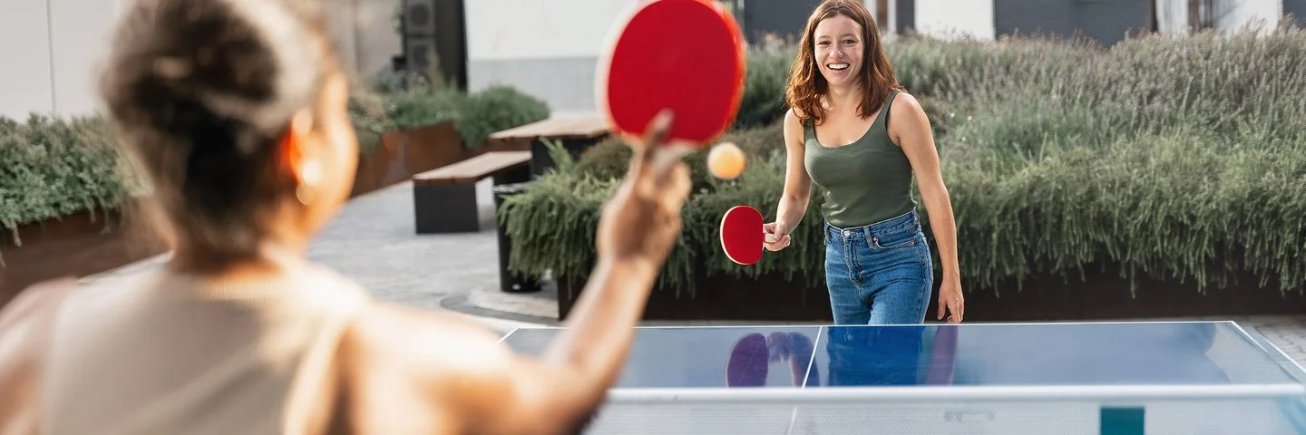Zwei junge Frauen in Freizeitkleidung spielen Tischtennis in einer öffentlichen Grünanlage.