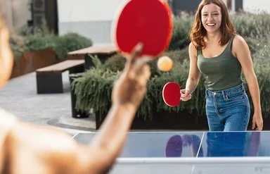 Zwei junge Frauen in Freizeitkleidung spielen Tischtennis in einer öffentlichen Grünanlage.