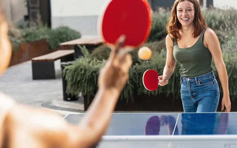 Zwei junge Frauen in Freizeitkleidung spielen Tischtennis in einer öffentlichen Grünanlage.