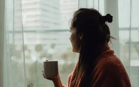 Frau mit Winterdepression schaut aus dem Fenster, sie hält dabei eine Tasse in der Hand.