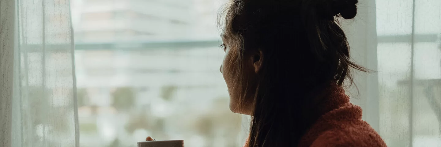 Frau mit Winterdepression schaut aus dem Fenster, sie hält dabei eine Tasse in der Hand.