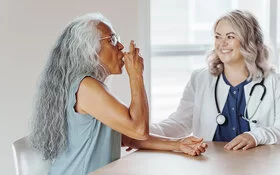 Eine Frau mit Asthma benutzt Asthmaspray. Eine Ärztin steht neben ihr und erklärt ihr die Anwendung.