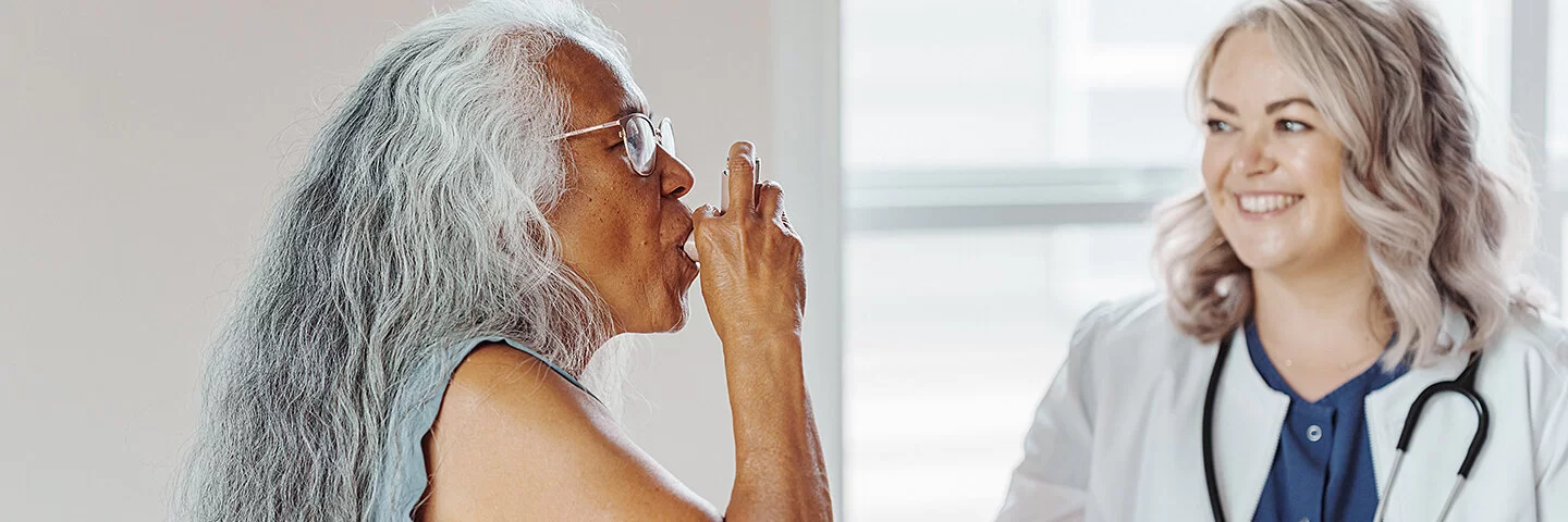 Eine Frau mit Asthma benutzt Asthmaspray. Eine Ärztin steht neben ihr und erklärt ihr die Anwendung.