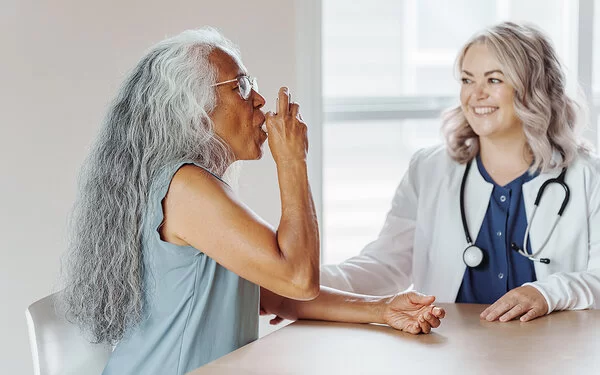 Eine Frau mit Asthma benutzt Asthmaspray. Eine Ärztin steht neben ihr und erklärt ihr die Anwendung.