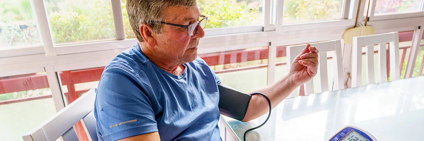 Ein Mann mit Übergewicht sitzt an einem Tisch und misst seinen Blutdruck.