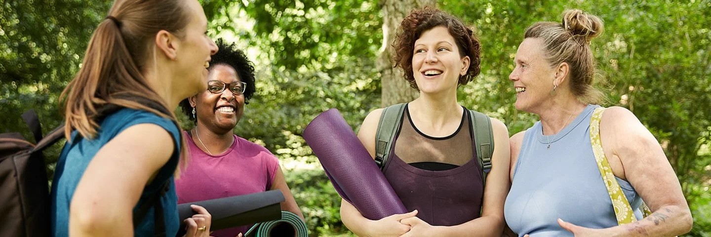 Vier Frauen stehen im Grünen und unterhalten sich. Sie tragen sportliche Kleidung und haben Yogamatten oder Sporttaschen dabei. Im Hintergrund sind Bäume und dichtes Blattwerk zu sehen. Das Licht wirkt natürlich und sommerlich.
