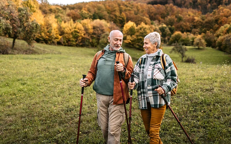 Ein älters Paar wandert mit Nordic-Walking-Stöcken durch eine herbstliche Landschaft.