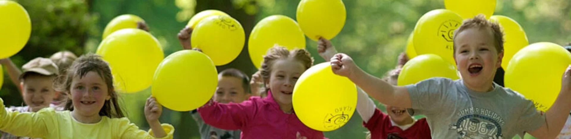 Lachende Kinder rennen bei Sonnenschein mit gelben Luftballons in der Hand durch einen Wald.