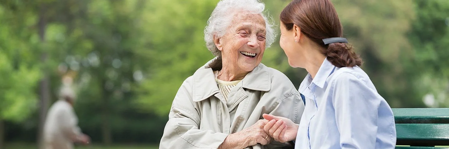 Eine ältere und eine jüngere Frau sitzen zusammen auf einer Parkbank. Sie lachen und unterhalten sich miteinander. Die jüngere Frau hat ein aufgeschlagenes Buch in der Hand. Im Hintergrund ist ein Mann mit einem Rollator zu sehen.