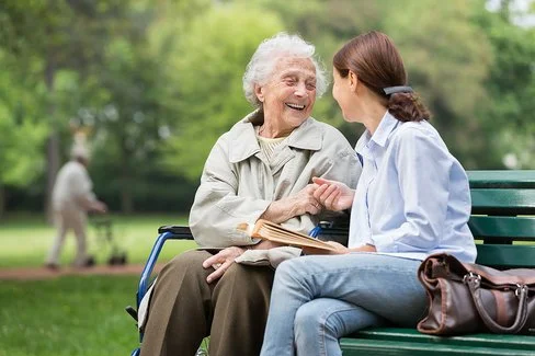 Eine ältere und eine jüngere Frau sitzen zusammen auf einer Parkbank. Sie lachen und unterhalten sich miteinander. Die jüngere Frau hat ein aufgeschlagenes Buch in der Hand. Im Hintergrund ist ein Mann mit einem Rollator zu sehen.