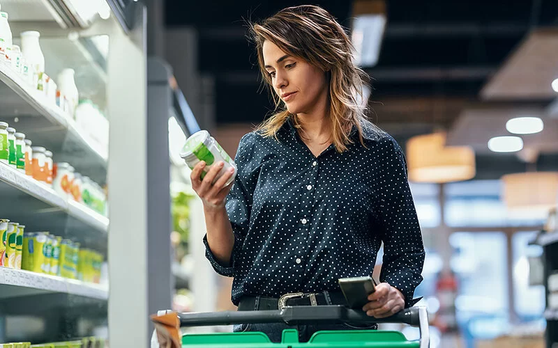 Eine junge Frau ist im Supermarkt einkaufen. Sie hält ein Glas in der Hand und schaut sich an, welche Zutaten enthalten sind.
