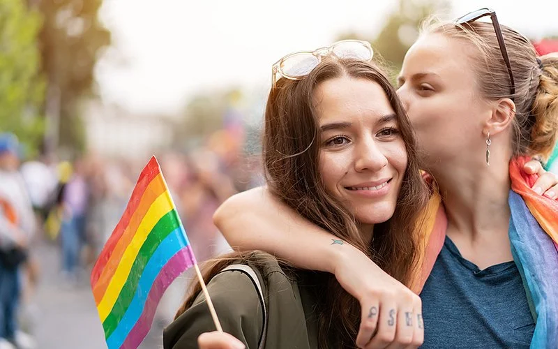 Zwei jüngere Frauen sind auf einem Pride-Event. Die Frau links ist von vorne zu sehen, sie schwenkt mit der rechten Hand eine kleine Regenbogenflagge aus Papier, die Frau rechts ist von der Seite zu sehen und küsst die andere Frau.