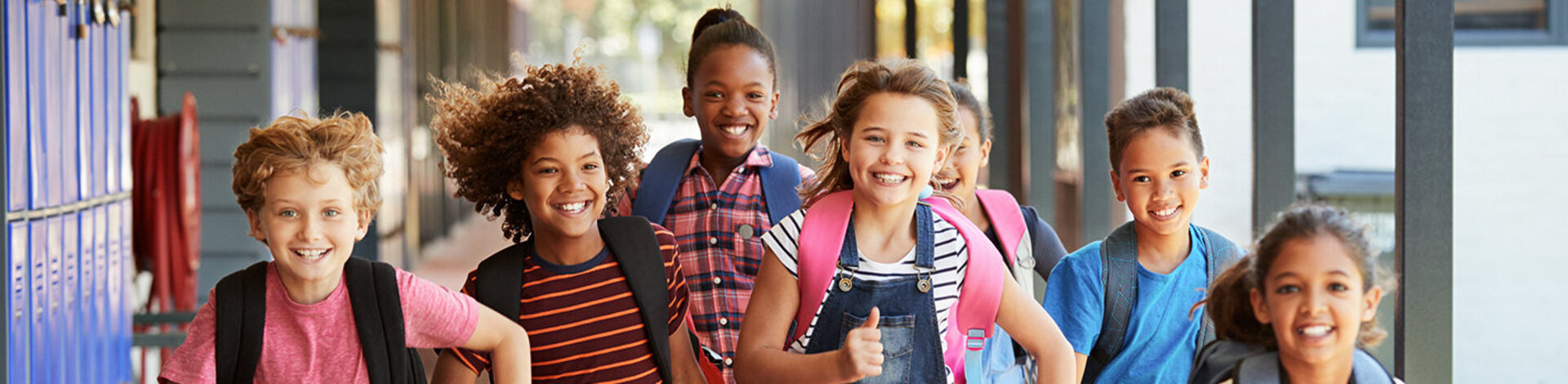 Sieben junge Schüler, vier Mädchen, drei Jungs- mit unterschiedlichen Hautfarben, bunt gekleidet, rennen strahlend mit ihren Schulrucksäcken durch eine Schule.