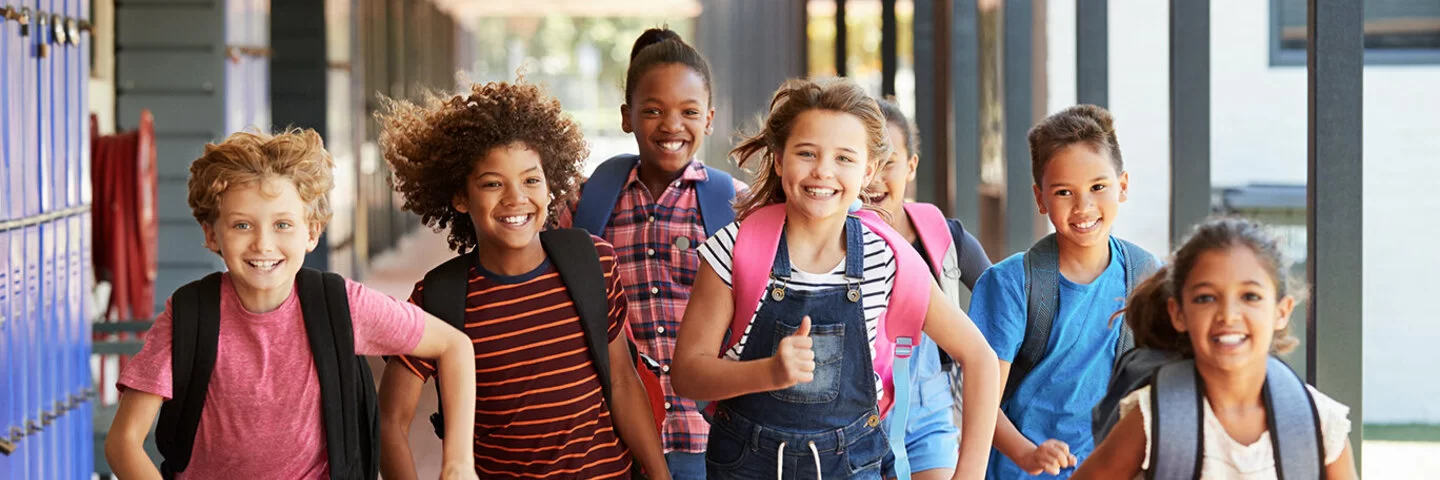 Sieben junge Schüler, vier Mädchen, drei Jungs- mit unterschiedlichen Hautfarben, bunt gekleidet, rennen strahlend mit ihren Schulrucksäcken durch eine Schule.
