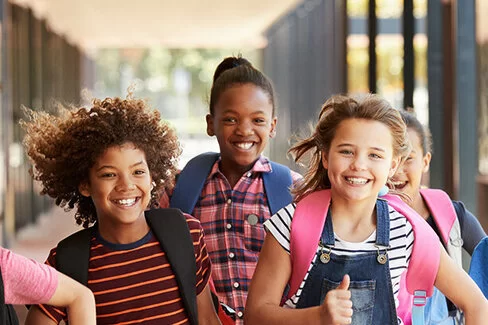 Sieben junge Schüler, vier Mädchen, drei Jungs- mit unterschiedlichen Hautfarben, bunt gekleidet, rennen strahlend mit ihren Schulrucksäcken durch eine Schule.