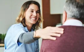 Eine freundlich lächelnde Pflegefachfrau sieht einen älteren Mann an und legt ihm die Hand auf die Schulter.