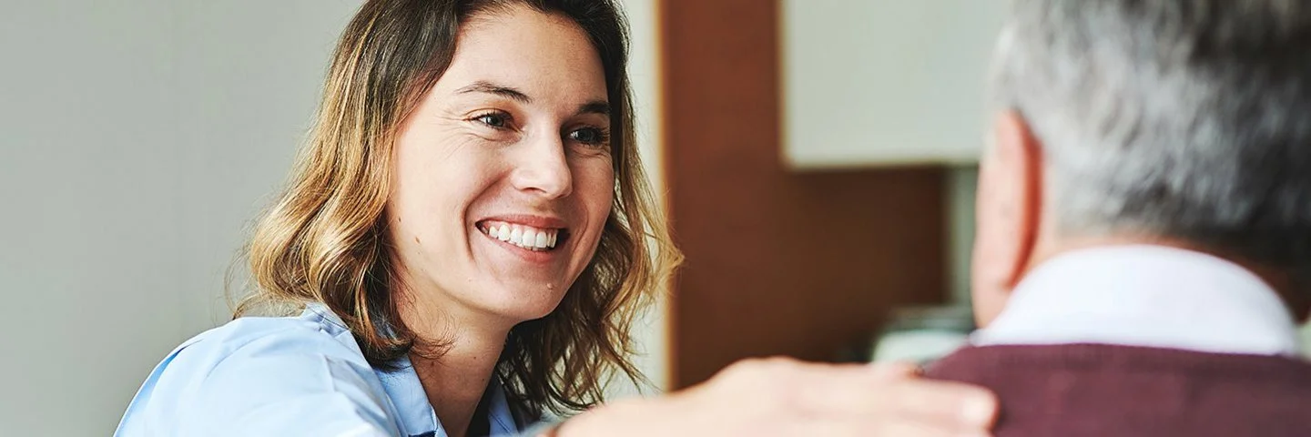 Eine freundlich lächelnde Pflegefachfrau sieht einen älteren Mann an und legt ihm die Hand auf die Schulter.