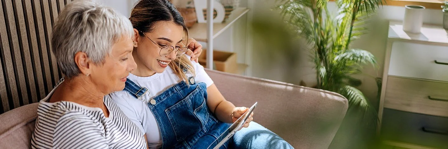Eine Seniorin und eine jüngere Frau sitzen auf einer Couch und blicken auf ein Tablet.