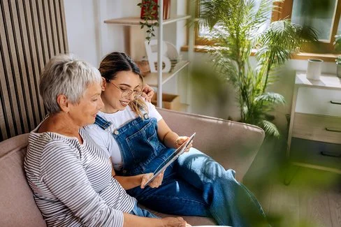 Eine Seniorin und eine jüngere Frau sitzen auf einer Couch und blicken auf ein Tablet.