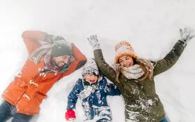Mutter, Vater und Sohn liegen mit dicker Winterkleidung im Schnee und lachen.