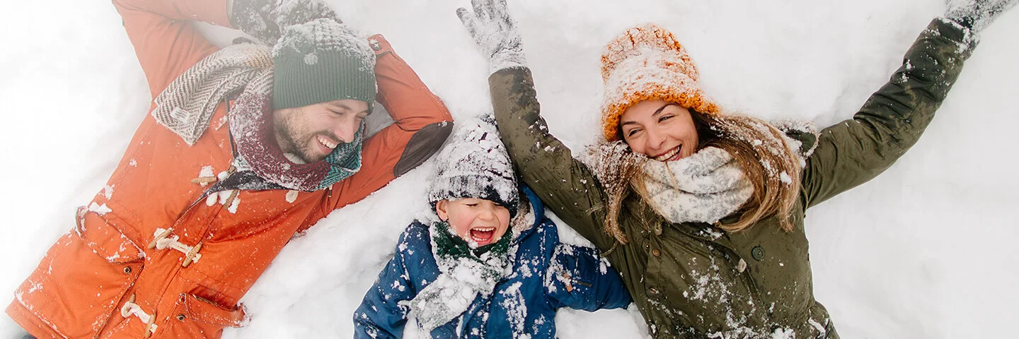 Mutter, Vater und Sohn liegen mit dicker Winterkleidung im Schnee und lachen.