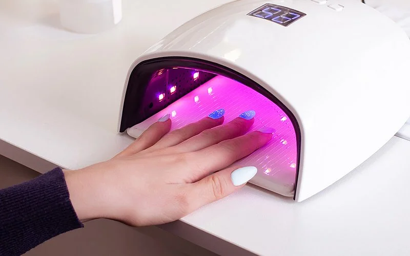 Eine Frau hält ihre Hand mit den blau und weiß lackierten Nägeln unter eine UV-Lampe.